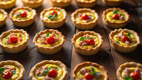 Twelve miniature quiches, each filled with spicy tuna salad and vibrant slices of ripe tomato.  The quiches are arranged on a rustic wooden surface, illuminated by soft, warm light, showcasing their golden-brown crusts and the colorful fill