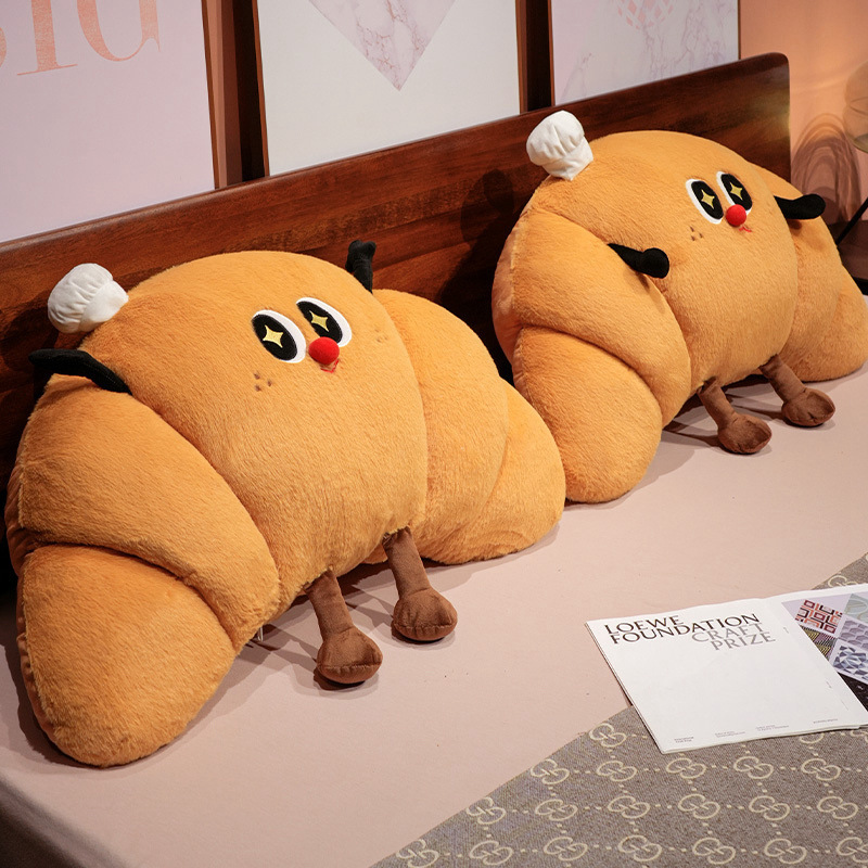 Two croissant plush pillows placed on a bed, soft croissant-shaped cushions with cute face design for cozy bedroom decor