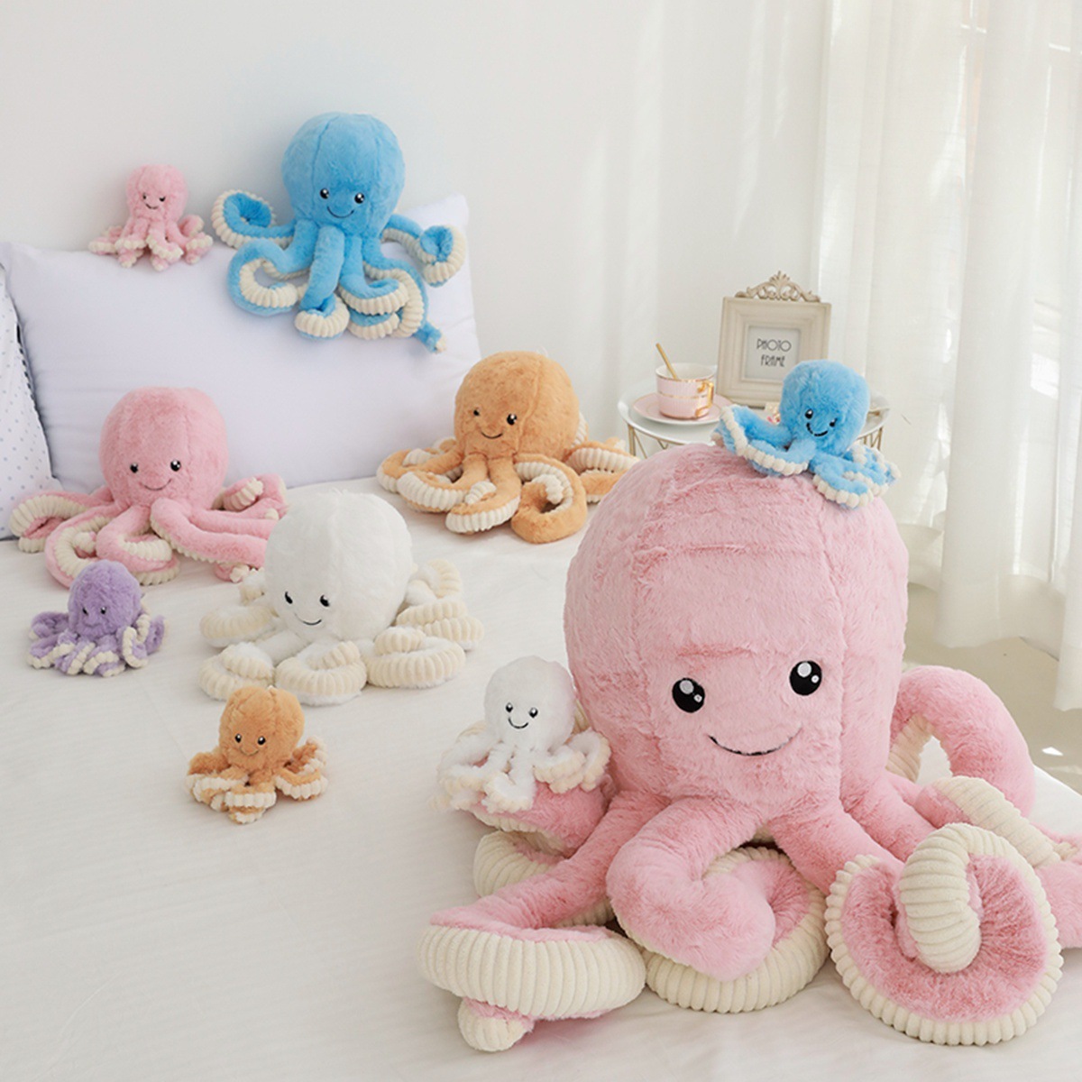 Octopus plush toy in multiple sizes and colors displayed on a bed, featuring soft fabric and long curly tentacles in a cozy home setting