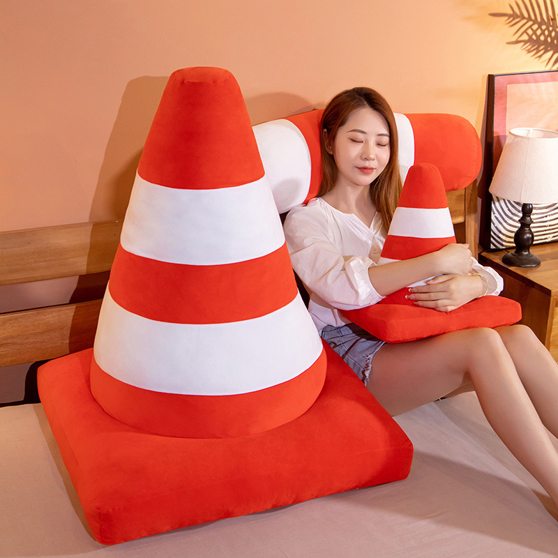 Traffic cone plush pillow with orange and white stripes used as a soft cushion on a bed, cozy plush design for hugging, lounging, or relaxing