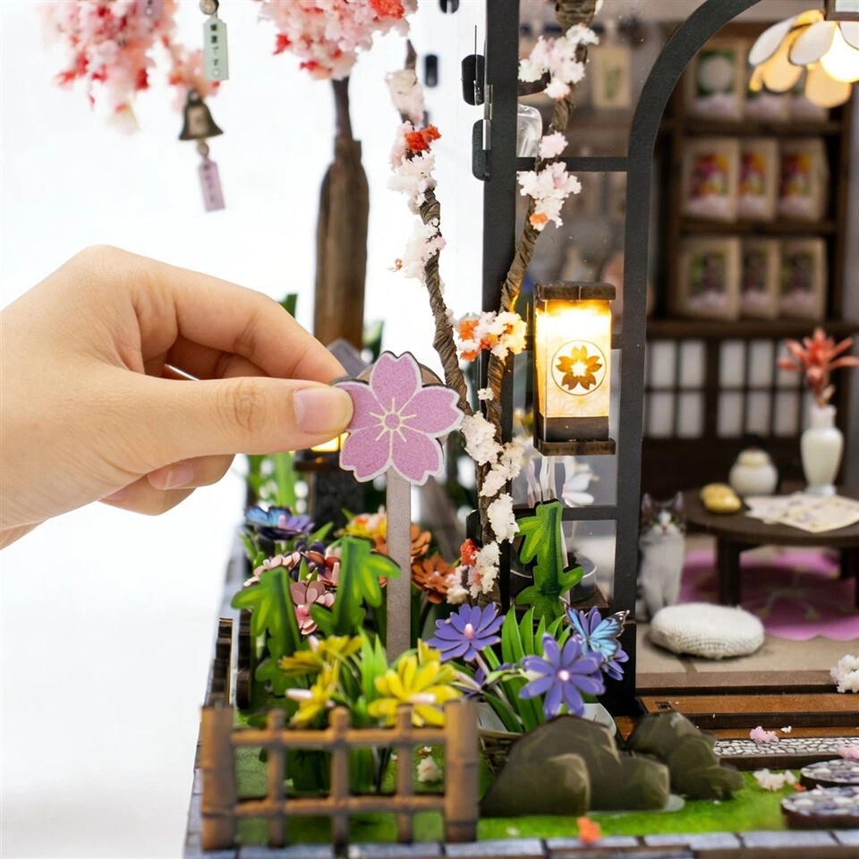 Close-up of Spring Sakura Greenhouse miniature house kit showing handcrafted flower garden details, warm LED lighting, and interactive DIY assembly scene