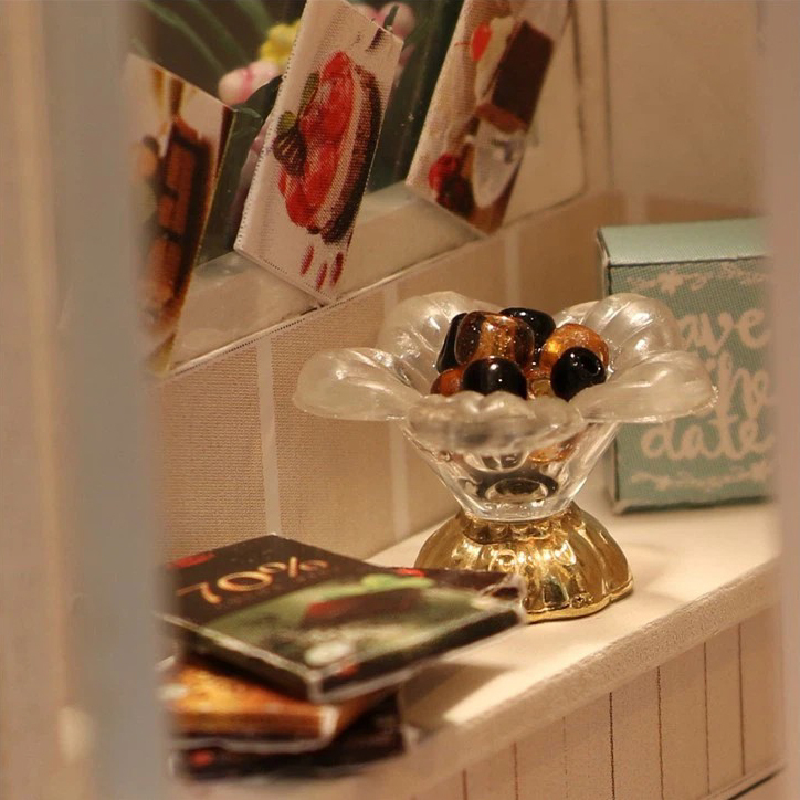 Miniature chocolate shop interior with decorative dessert bowl, printed menu cards, chocolate packaging, and soft warm lighting for cozy dollhouse display