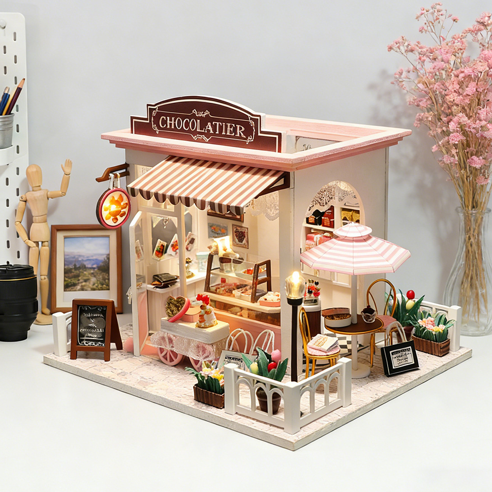 Side-angle view of handmade miniature chocolate boutique featuring outdoor café seating, striped umbrella, warm lighting, and detailed dessert shop accessories