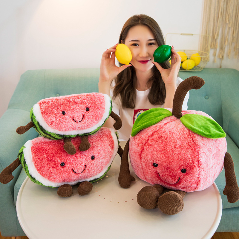 Smiling watermelon and cherry plush toys displayed together on a table, featuring soft plush fabric, embroidered faces, and playful dangling arms and legs in a cheerful home setting