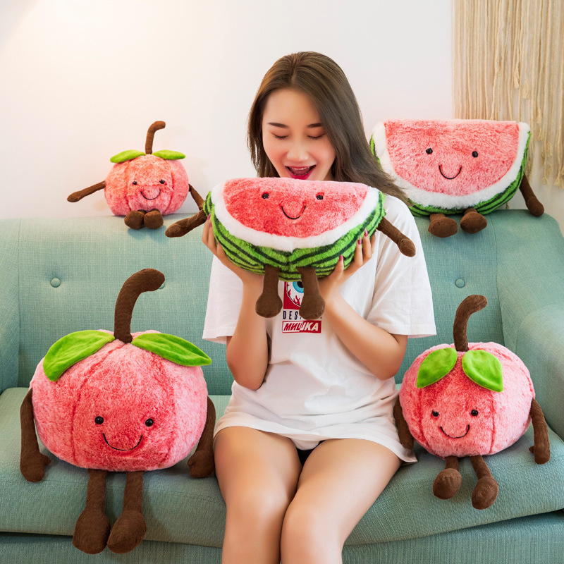 Watermelon and cherry plush toys displayed on a sofa, featuring smiling embroidered faces, soft plush fabric, and playful dangling arms and legs for cozy room decor and companionship