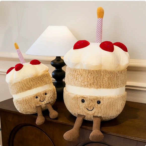 Birthday Cake Plush Toys displayed on a wooden cabinet beside a table lamp, featuring smiling faces, soft plush fabric, and charming cake-inspired design