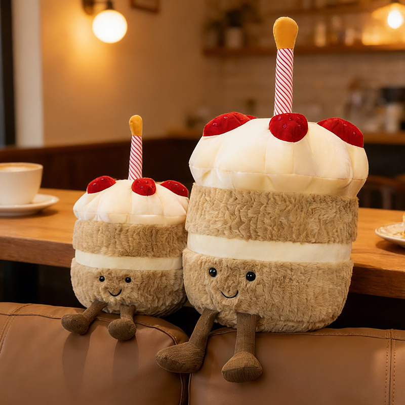 Birthday Cake Plush Toys in two sizes displayed in a cozy cafe setting, showcasing soft plush textures, layered cake details, and adorable dangling legs