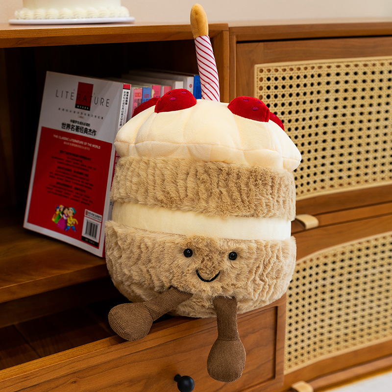 Birthday Cake Plush Toy displayed on a bookshelf, featuring a smiling face, fluffy cake-inspired design, soft plush fabric, and dangling legs for playful home decor
