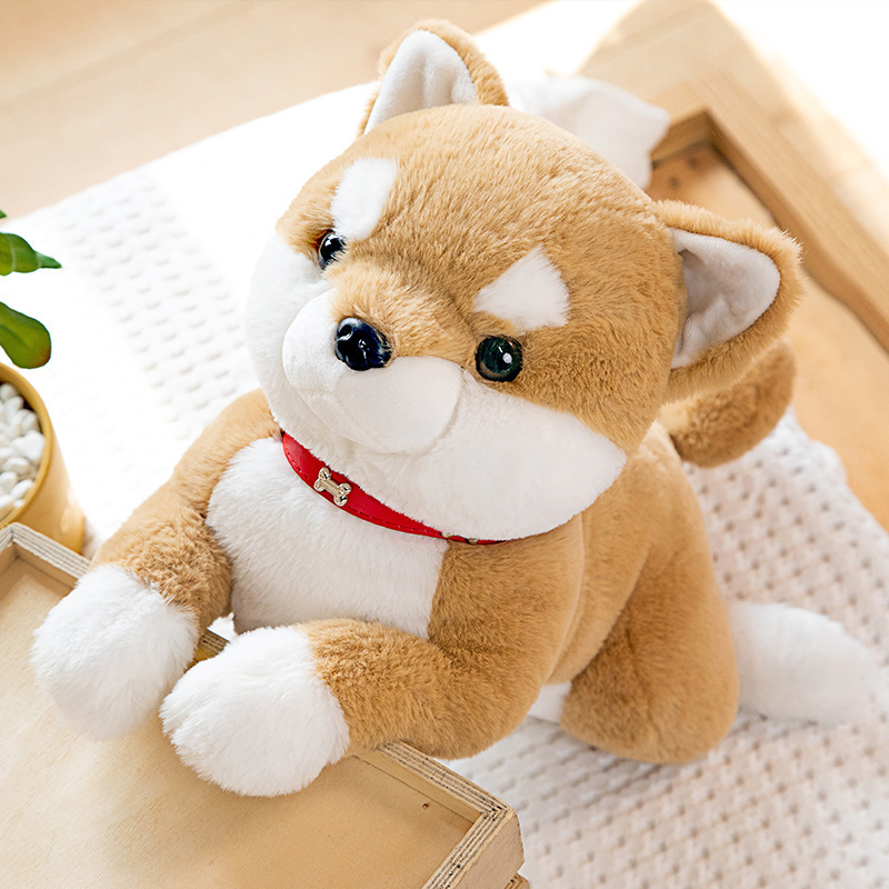 Brown Shiba Inu plush toy with a red collar and bell displayed on a wooden crate in a cozy room setting