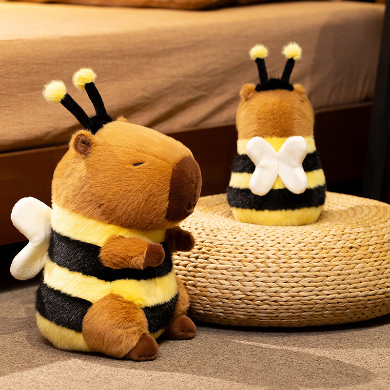 Bee Capybara Plush Toy showing front and back details, including plush wings, fuzzy antennae, and striped bee-inspired design, displayed on a woven cushion in a warm bedroom atmosphere