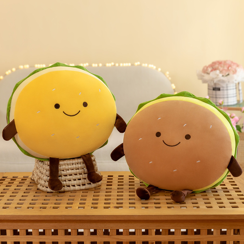 Cute burger plush pillow in yellow and brown color options displayed on a wooden bench, featuring smiling faces, soft plush fabric, and a cozy food-themed home decor style