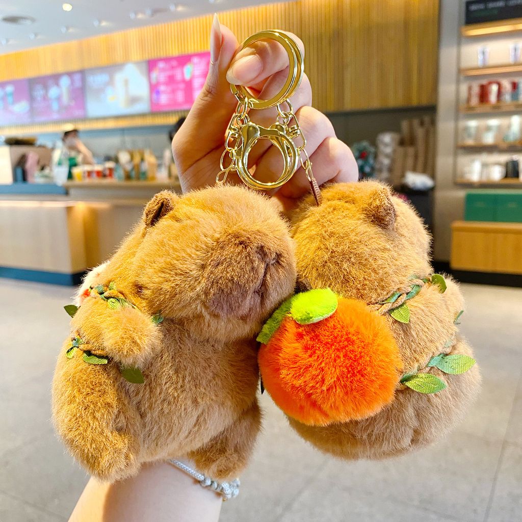 Two cute capybara plush keychain charms, one shown from the front and the other wearing an orange fruit backpack.