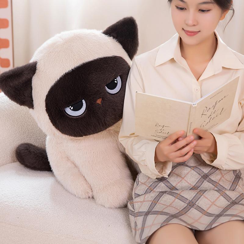 Sitting ragdoll cat plush toy beside a book on a sofa for cozy home decor and relaxation