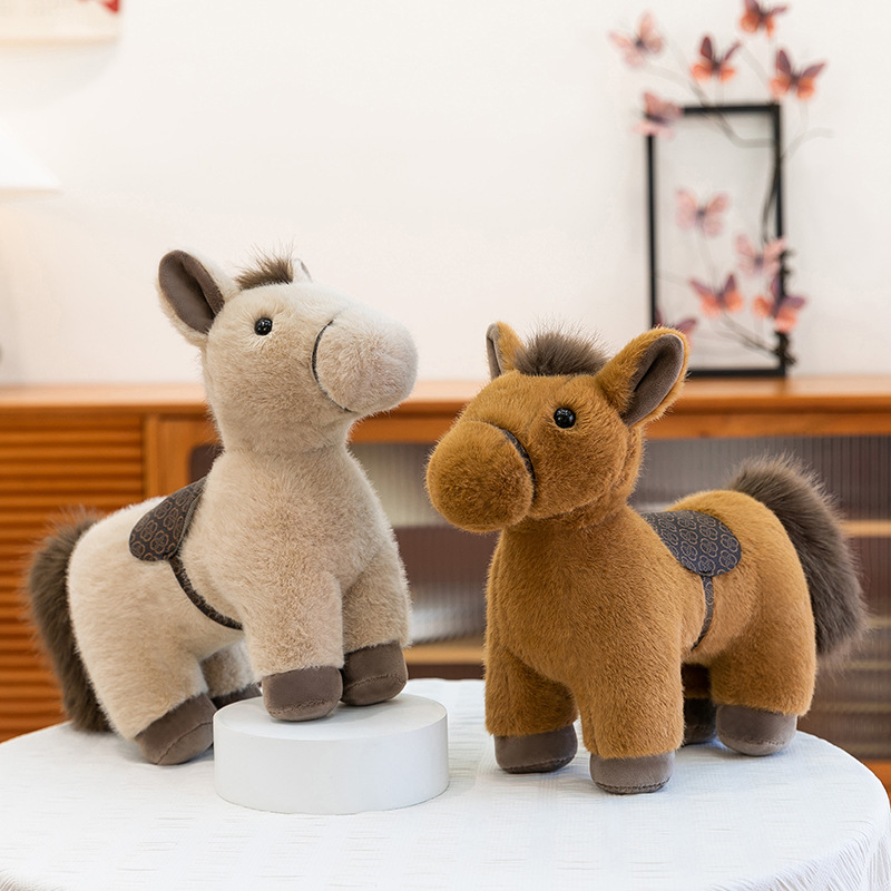 Realistic pony plush toy in light khaki color with soft fur and saddle, displayed on a tabletop in a warm home setting, showing a gentle standing pose, suitable for hugging, decorating shelves, or adding a cozy and calming atmosphere to the room