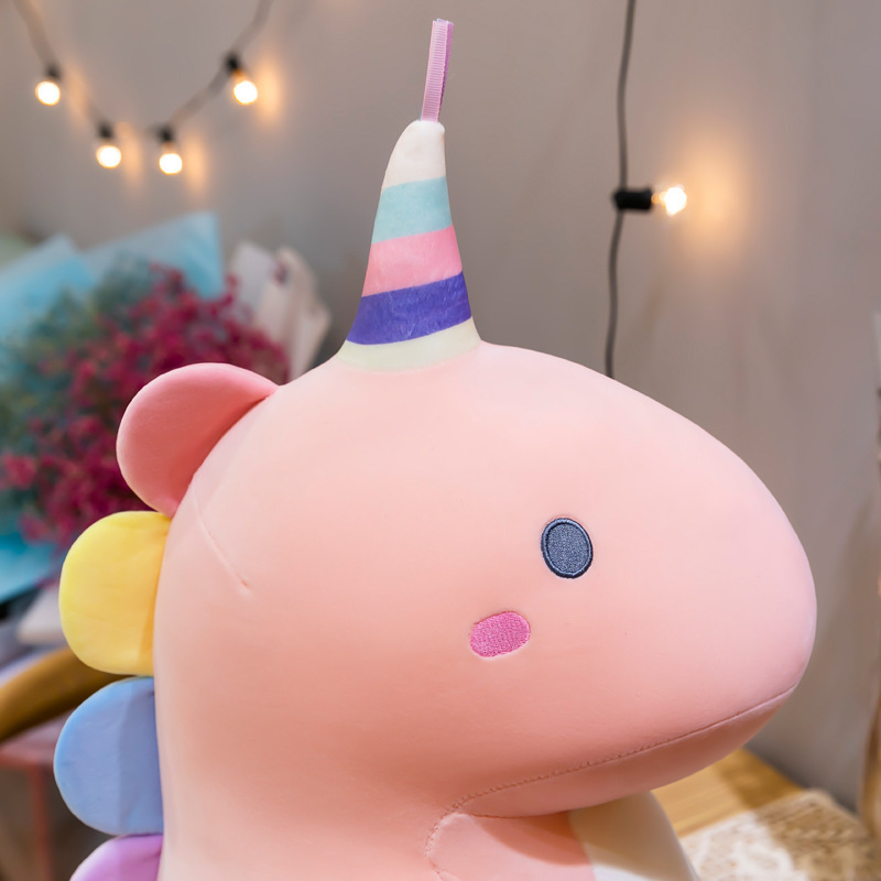 Candy dinosaur plush toy head detail with soft pastel fabric, embroidered facial features, and striped decorative horn, shown in a cozy indoor setting with warm lighting