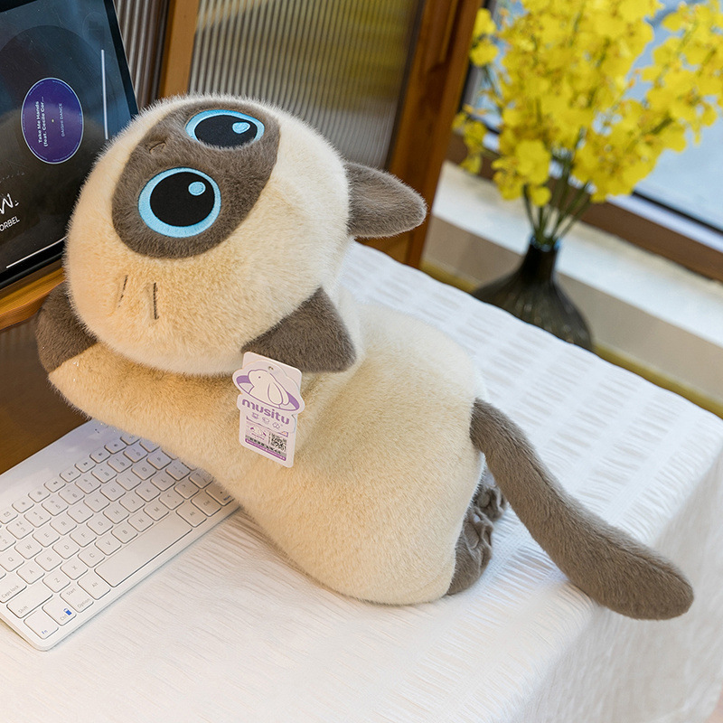 Siamese lying cat plush toy placed beside a keyboard, with a long relaxed body and plush fabric, adding warmth and comfort to a cozy home workspace or bedroom setting
