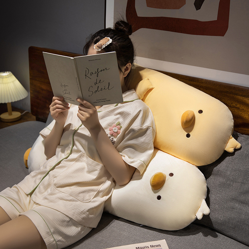 Milk chick plush pillow used as a backrest on bed, long chicken body pillow in milky yellow and milky white, soft plush fabric providing cozy support for relaxing and reading in bedroom