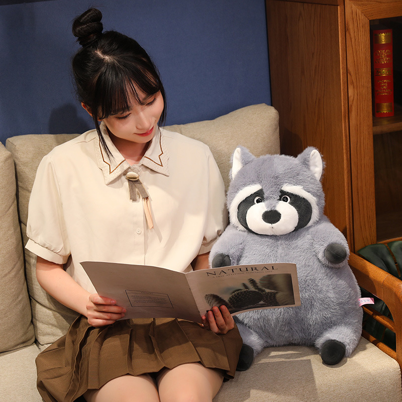 Chubby raccoon plush toy sitting on a sofa beside a book, featuring soft plush fabric and a round, huggable shape, creating a cozy and relaxing home atmosphere for reading and lounging