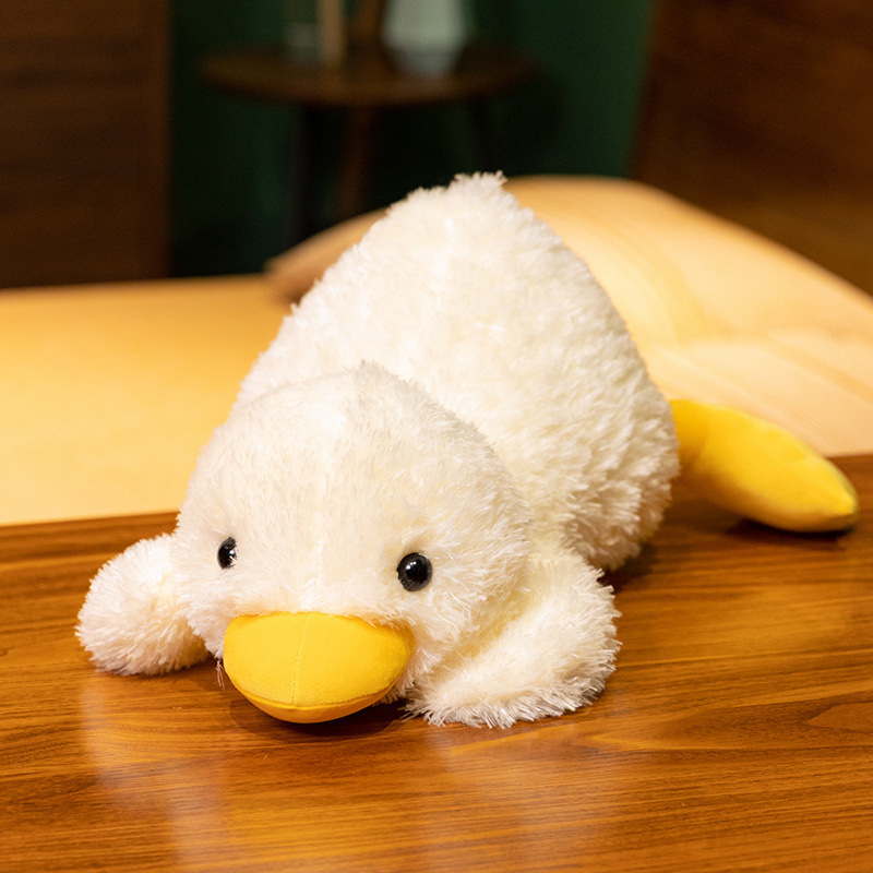 Lying white duck plush pillow resting on a wooden surface, featuring soft fluffy texture, rounded body, and yellow beak, designed for hugging, relaxing, and cozy home decor