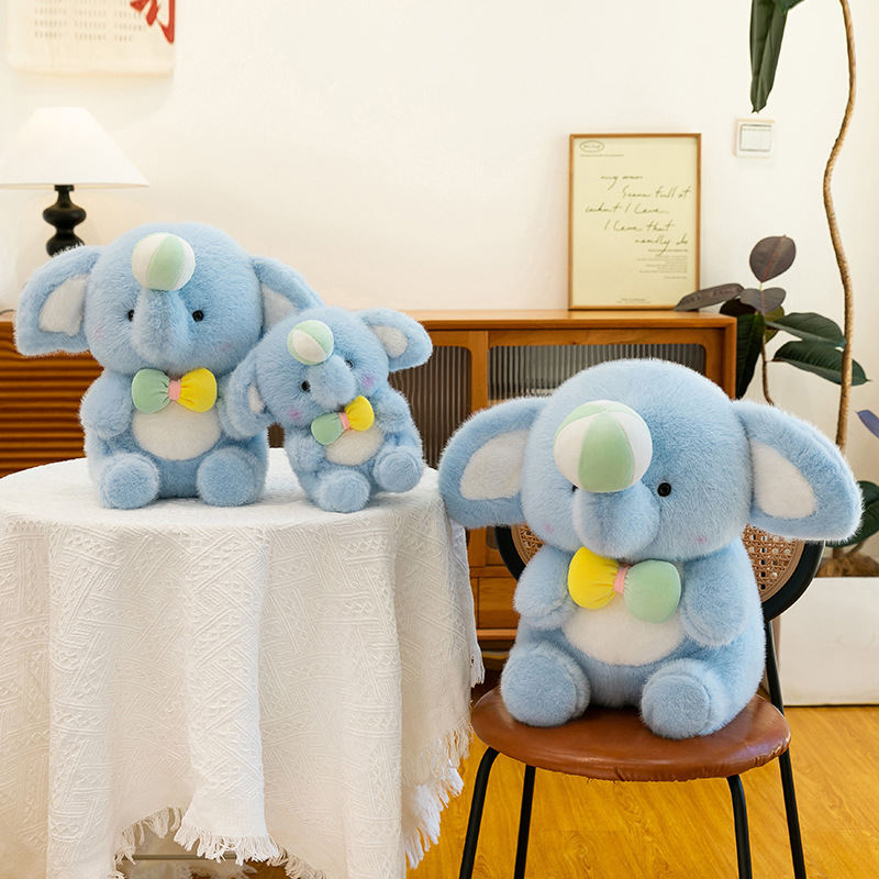 Happy elephant plush toys in multiple sizes displayed on a table and chair, showing size comparison, soft plush texture, pastel blue color, and a cozy home decor atmosphere
