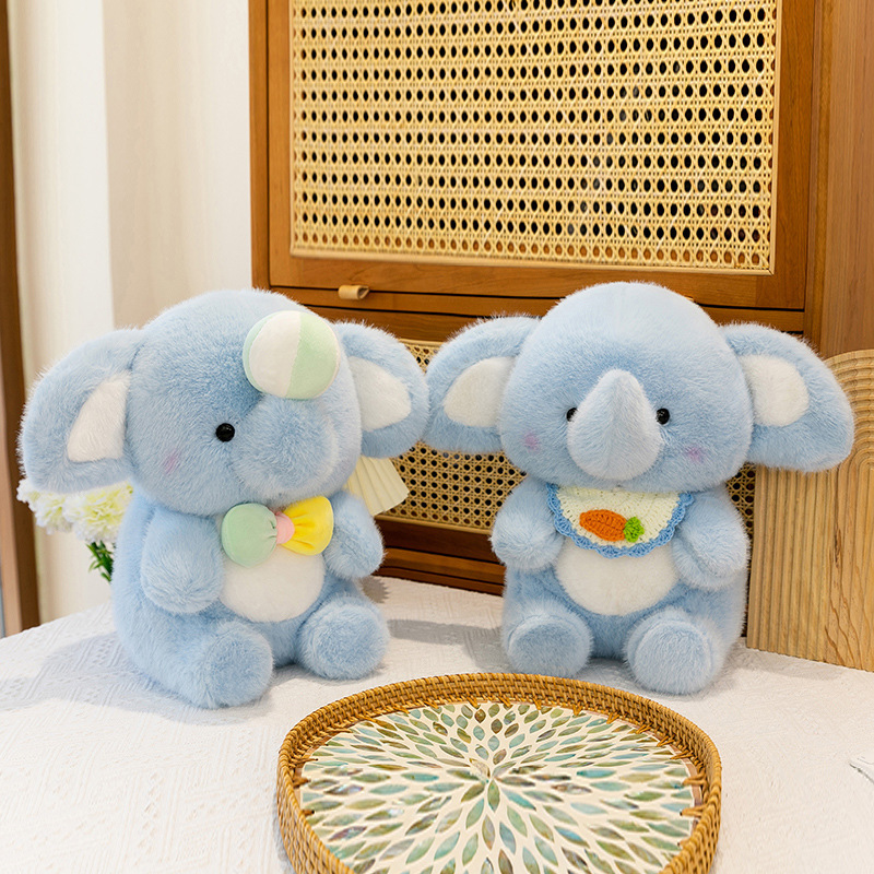 Happy elephant plush toys displayed on a table, featuring bib and bow variants with soft pastel blue plush fabric, shown as cute decorative companions for cozy home spaces