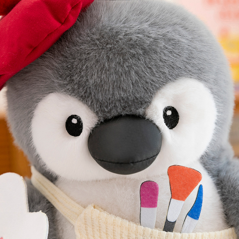 Close-up of painter penguin plush toy face with fluffy fabric and stitched details, featuring a cute apron with colorful brush design