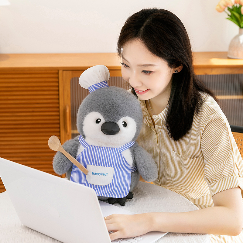 Chubby chef penguin plush toy placed on a table beside a laptop, showing a soft and comforting plush companion suitable for home, desk decor, or everyday relaxation