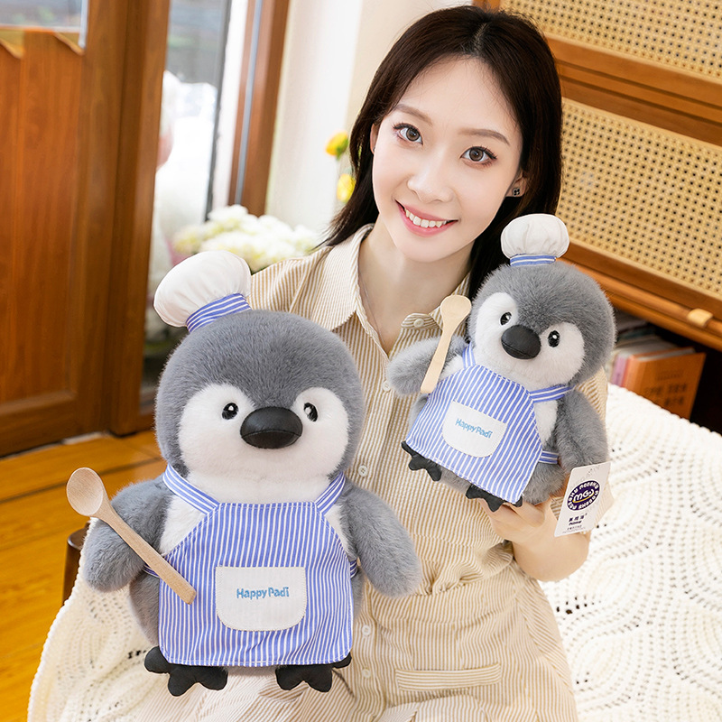 Chubby penguin plush toys in two sizes, wearing blue chef aprons and hats, shown together to highlight size comparison and soft, huggable texture in a cozy indoor setting