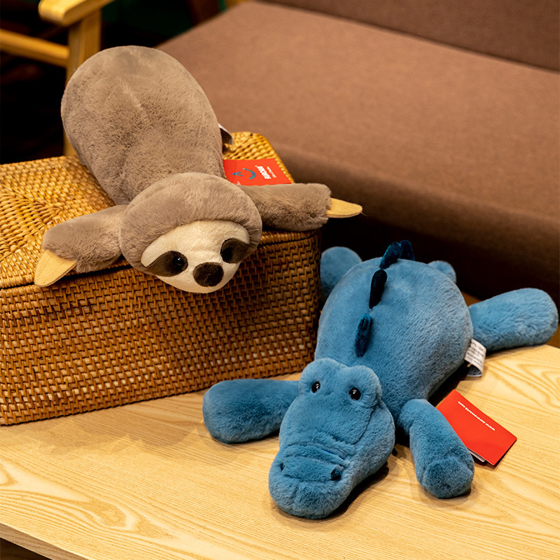 Lazy lying sloth and crocodile plush toys displayed on woven stool and wooden floor, soft stuffed animals for hugging and cozy home decor