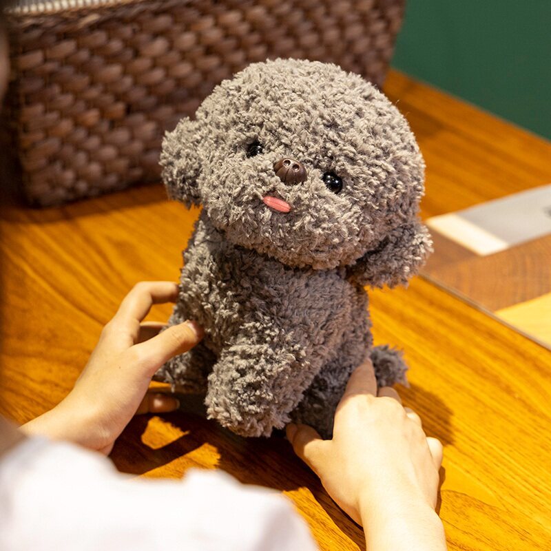 Gray Bichon Frise plush toy with tongue out, made of teddy plush fabric, displayed on a wooden table in a cozy indoor setting, showing soft texture and compact size