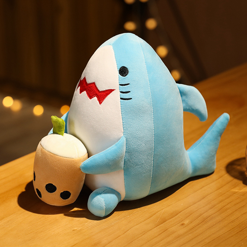 Shark milk tea animal plush toy displayed on a wooden table, soft blue plush fabric with rounded bubble tea cup detail in warm ambient lighting