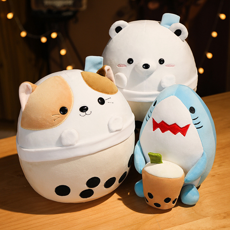 Milk tea animal plush toys featuring bear, cat and shark designs displayed together on a wooden table, soft rounded shapes with bubble tea cup details in a warm cozy setting