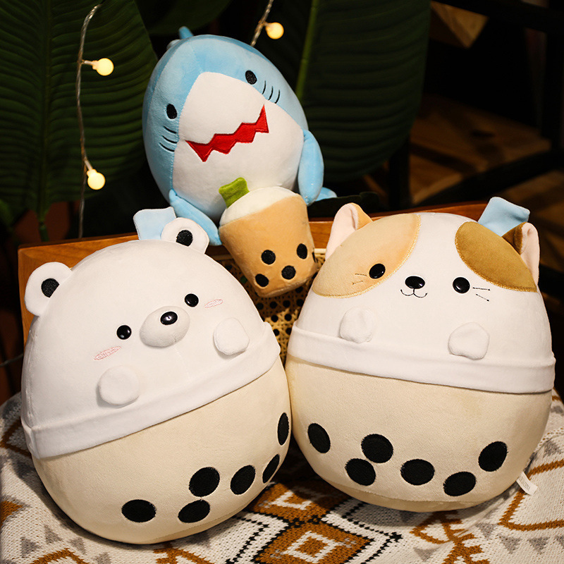 Milk tea animal plush toys featuring white bear, cat and shark designs displayed on a patterned table surface, with the shark leaning against a chair in a warm indoor setting