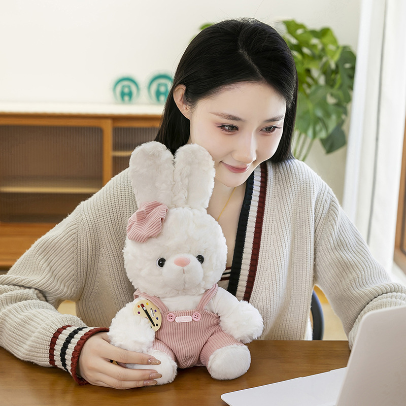 Mora bunny plush toy in pink corduroy overalls placed on a desk beside a laptop, soft stuffed animal for cozy home decor