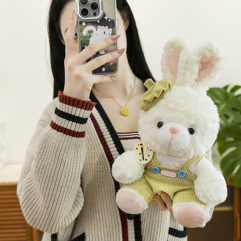 Mora bunny plush toy in green corduroy overalls held for size reference, soft texture and cozy indoor setting