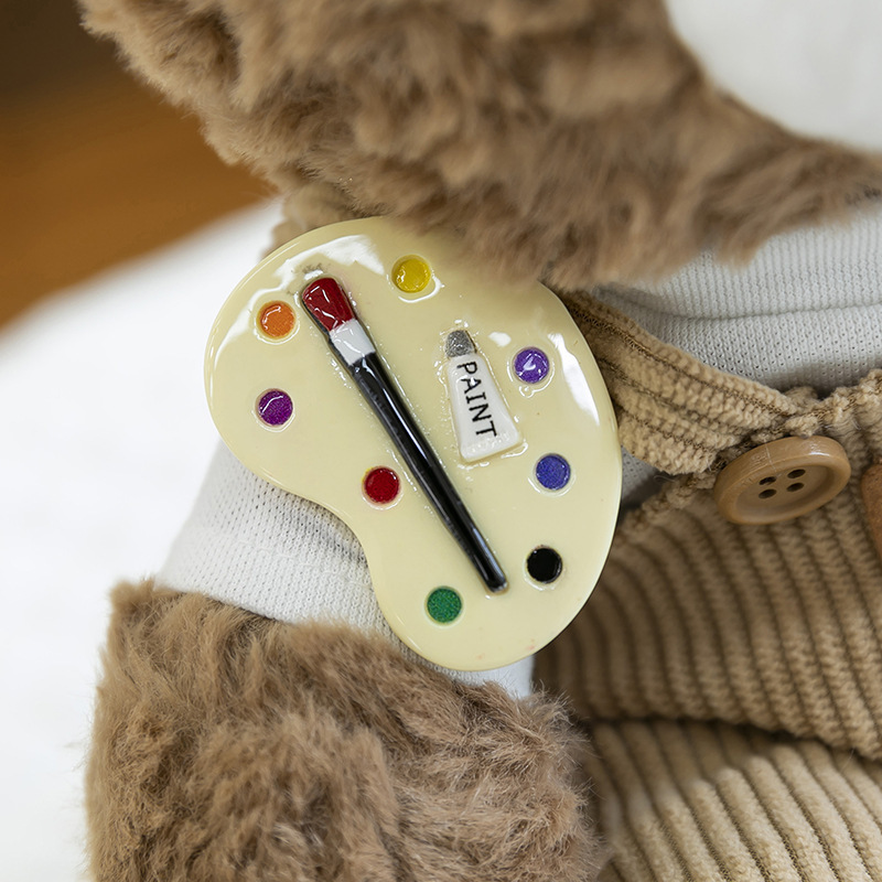 Artist palette badge detail on mocha bear plush toy, colorful paint design attached to corduroy overalls
