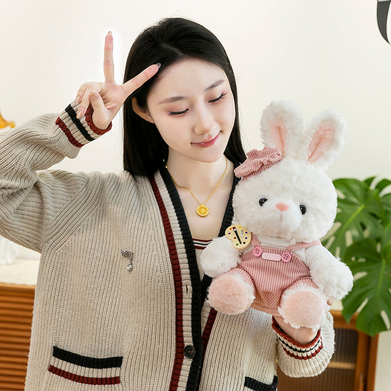 Mora bunny plush toy in pink corduroy overalls held for display, showing fluffy texture, soft pastel tones and artist palette badge detail