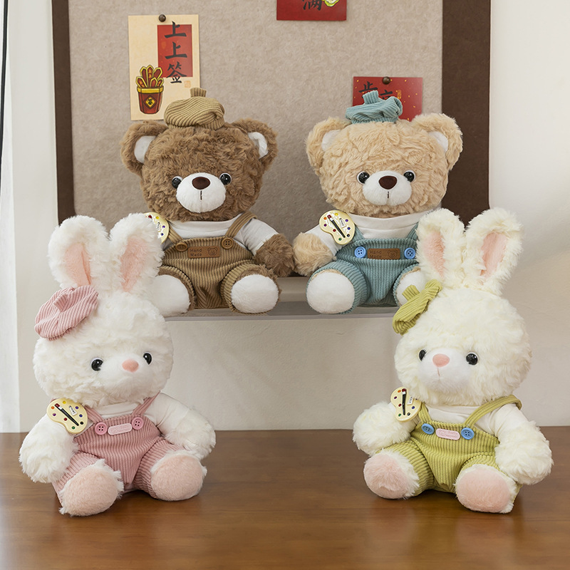 Artist mocha bear and mora bunny plush toys in pastel overalls displayed on a wooden table, cozy indoor setting with soft decorative background
