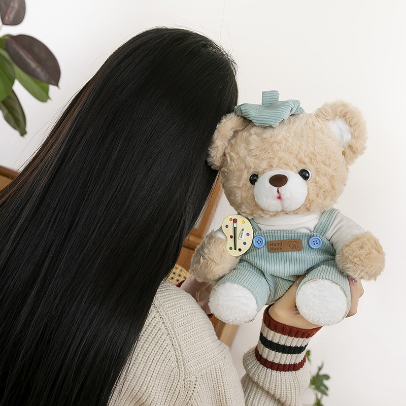 Mocha bear plush toy in blue corduroy overalls held up for display, fluffy fabric and artist palette badge detail