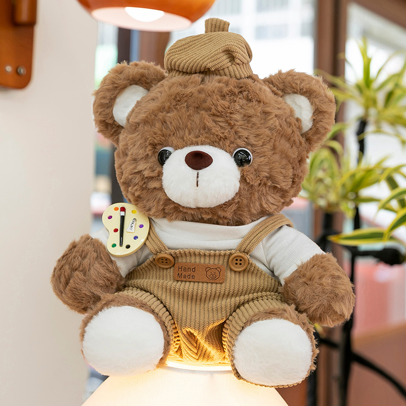 Mocha bear plush toy in brown corduroy overalls sitting on a lit lamp base, warm ambient lighting and cozy indoor decor setting