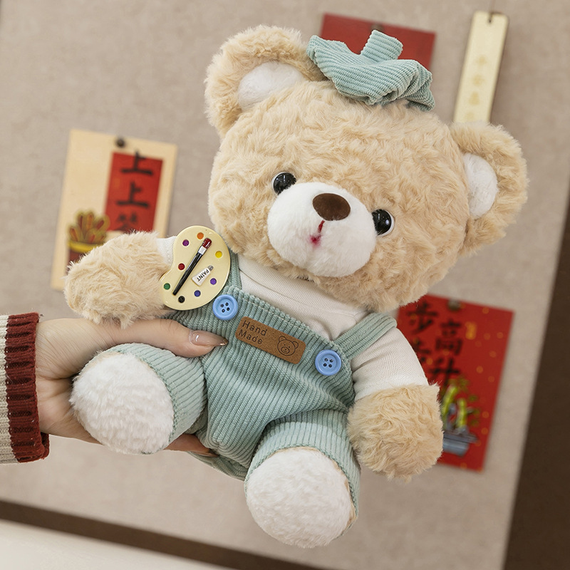 Mocha bear plush toy in blue overalls shown up close, fluffy plush fabric and detailed artist badge design