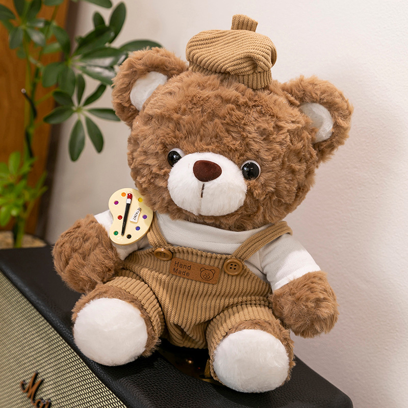 Mocha bear plush toy in brown corduroy overalls sitting on a speaker, soft stuffed animal with artist palette badge in cozy home setting