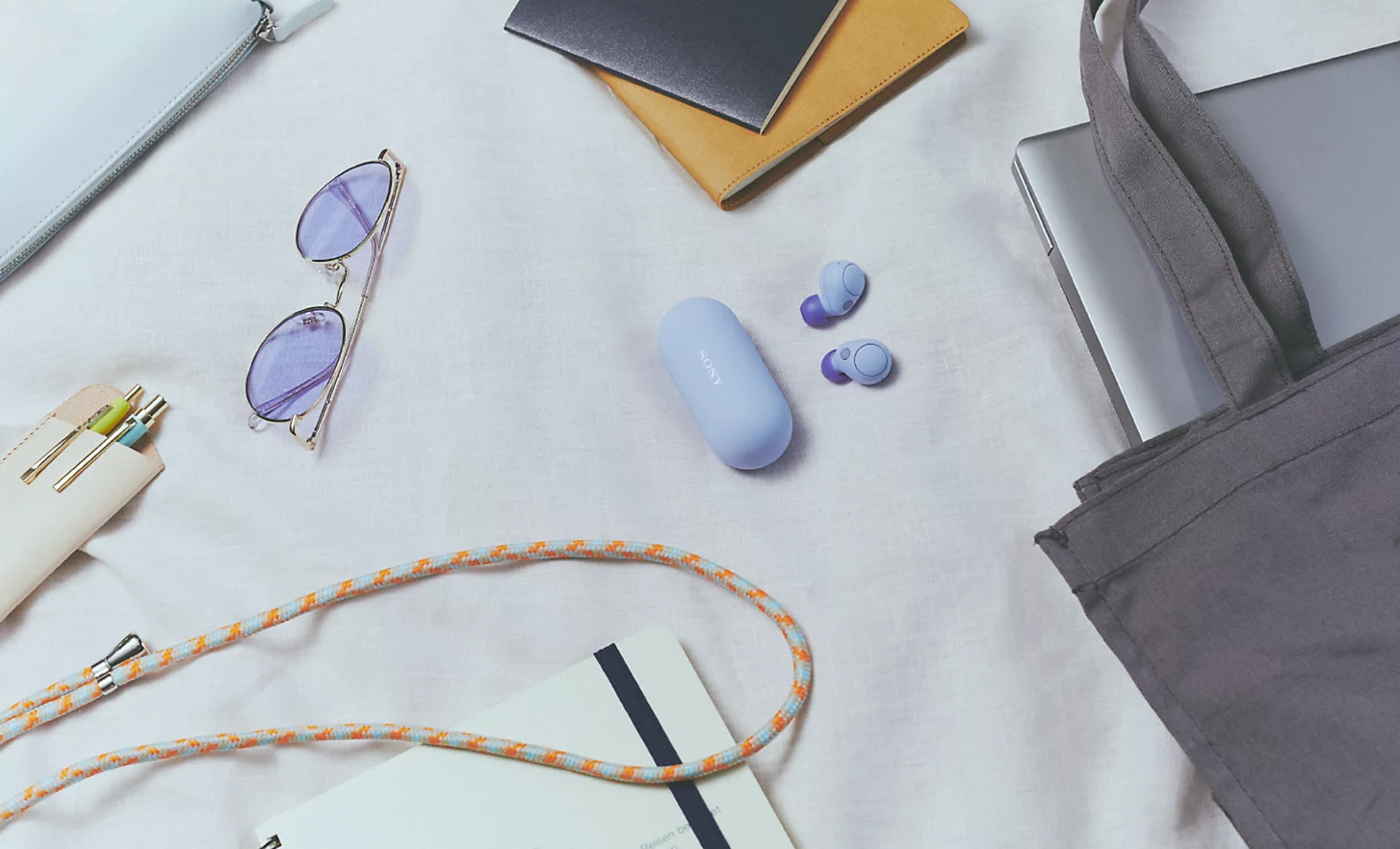 Image of the lavender WF-C700N headphones and case surrounded by various everyday items such as a laptop, bag and sunglasses
