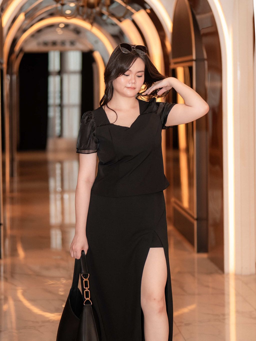 Plus-size fashion model adjusting her hair while wearing black sheer puff sleeve top and front-slit skirt.