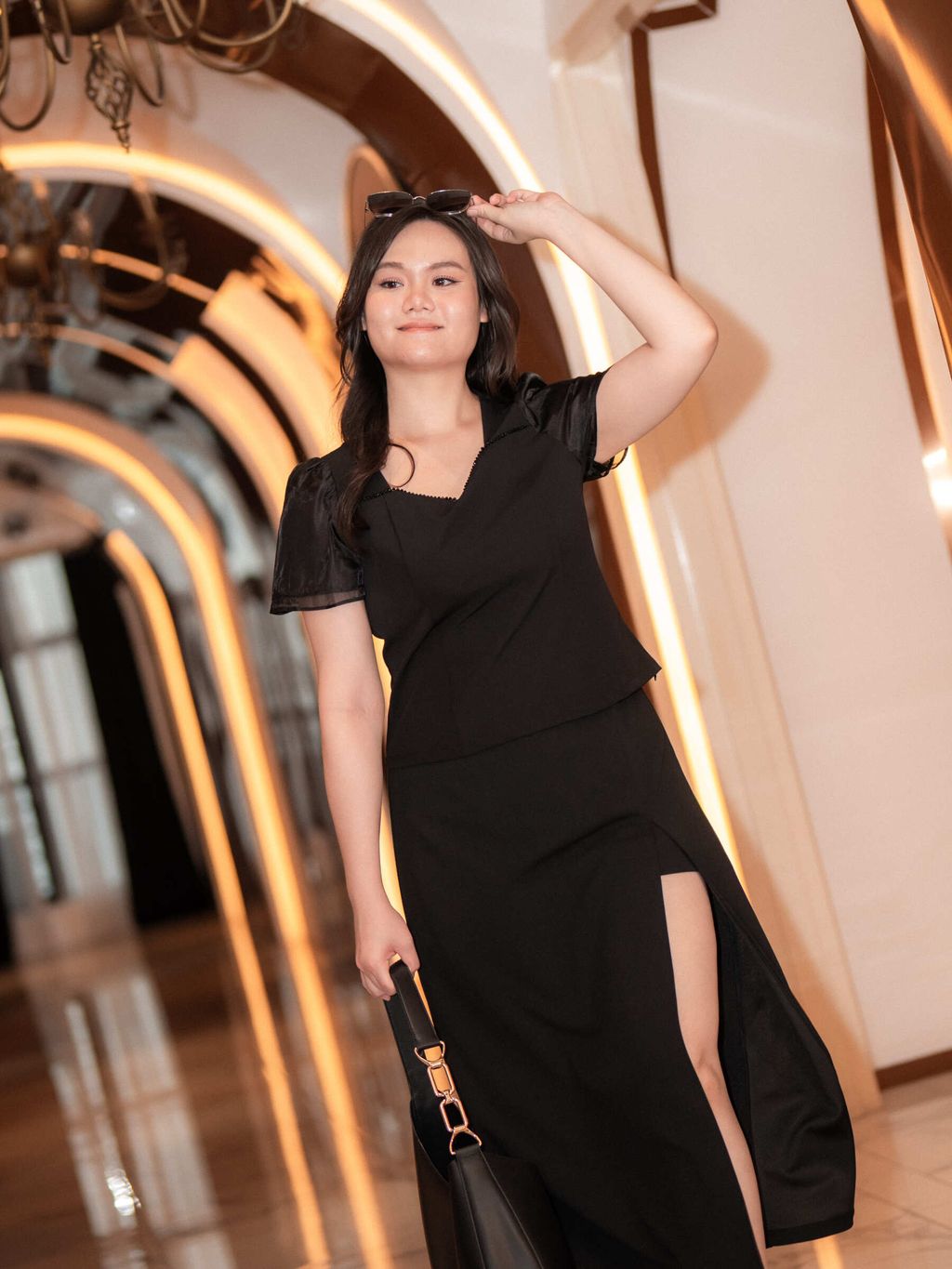 Confident plus-size woman walking in a luxurious hallway, styled in twist-neck black blouse and front-slit midi skirt.