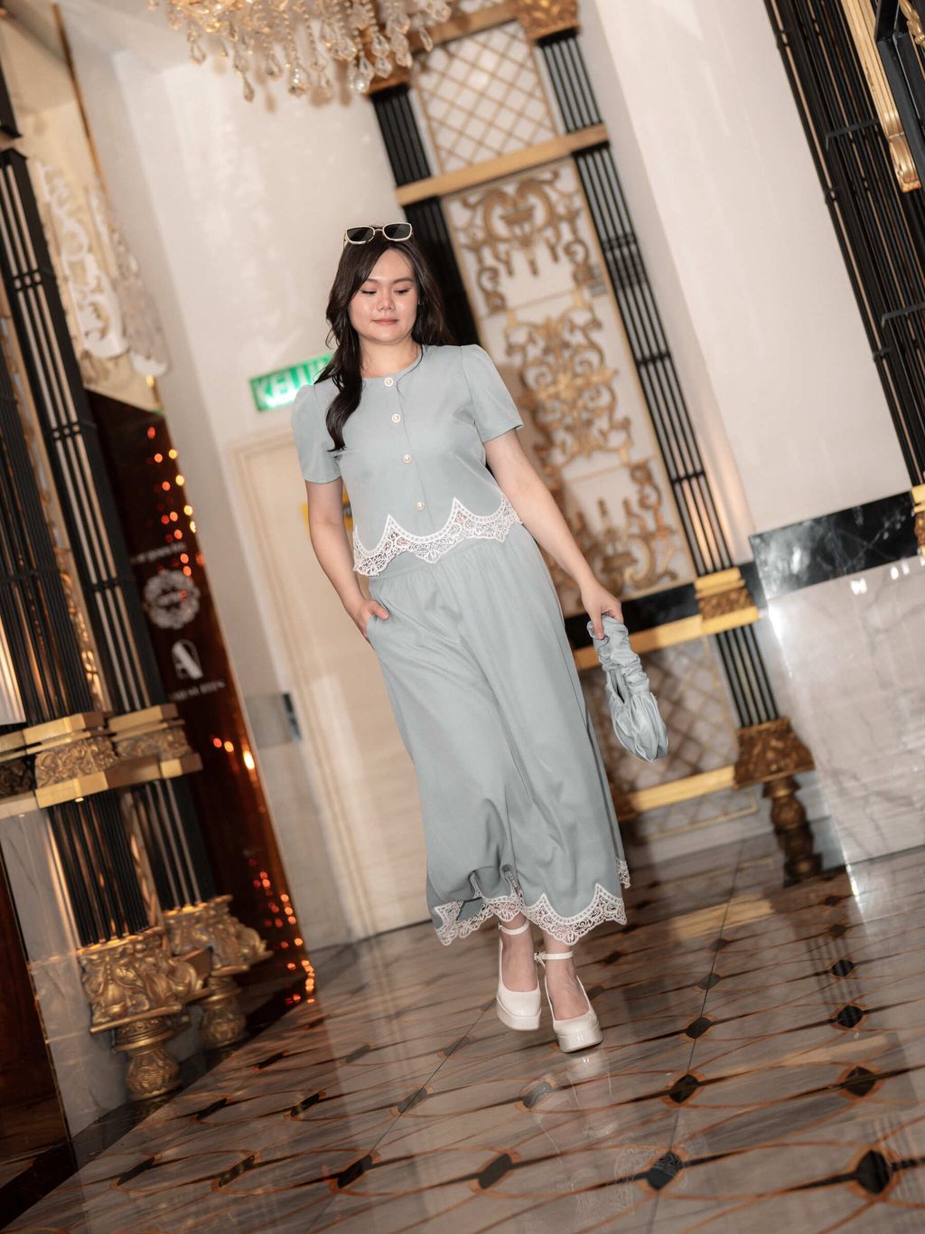Plus-size model walking confidently in a pale blue lace-trimmed top and skirt set in a grand hallway.