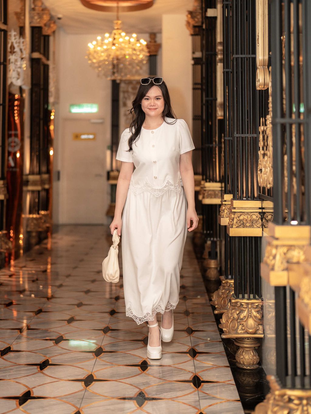 Model in white version of lace-trimmed set walking under a chandelier, exuding elegant charm.