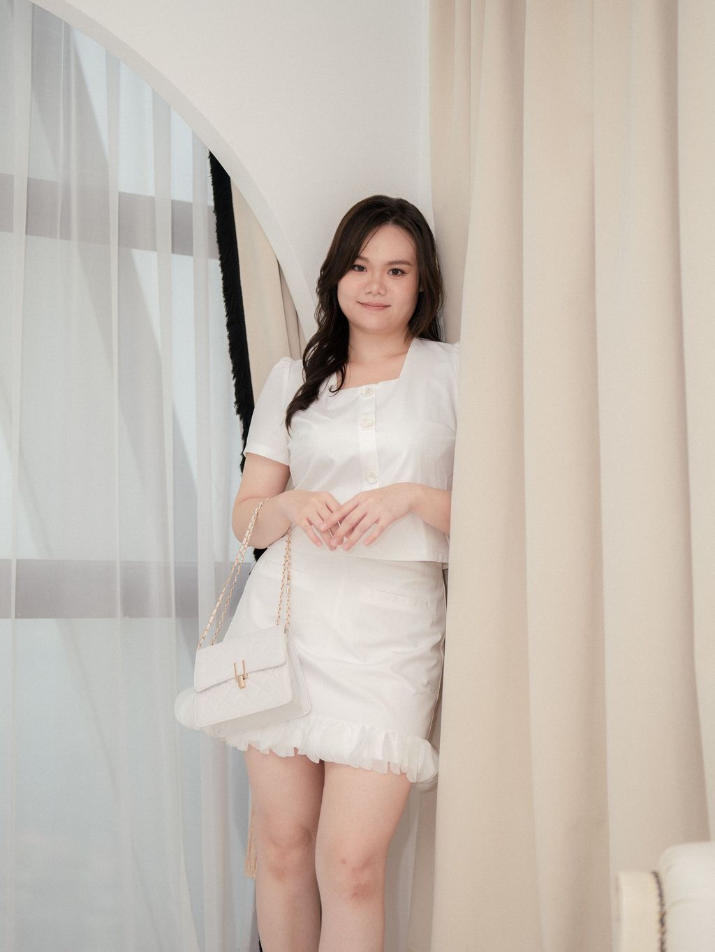 Plus-size model wearing a white blouse with floral pearl buttons, leaning gently by the curtain.