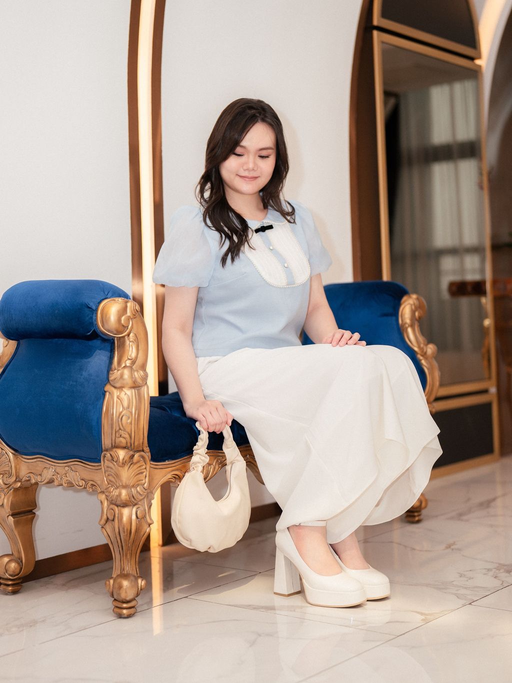 Plus-size model sitting gracefully in an ornate chair, wearing a puff sleeve blouse and holding a ruched cream handbag.