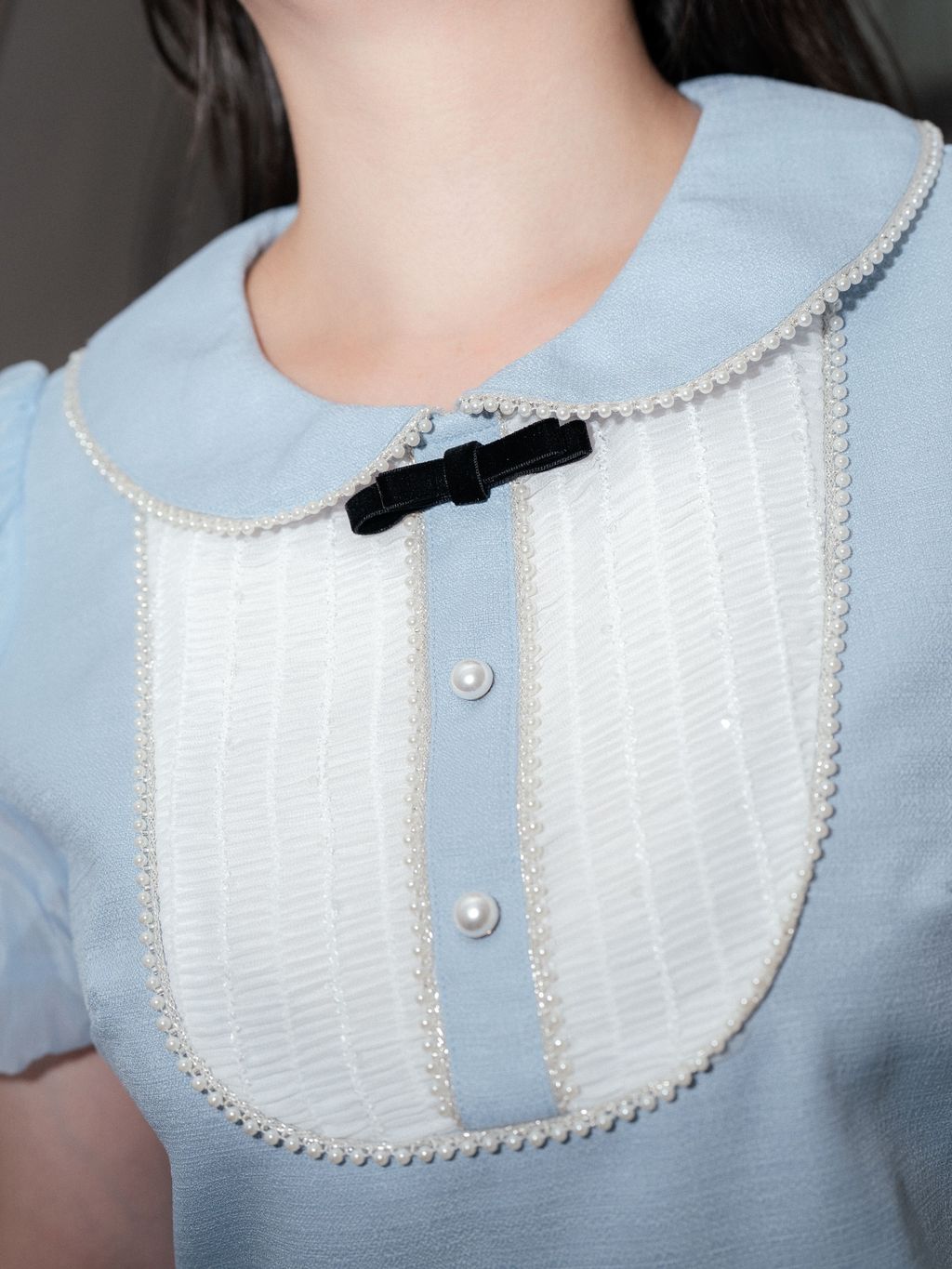 Close-up of bib detail with pearl-trim, pleated texture, and velvet bow.